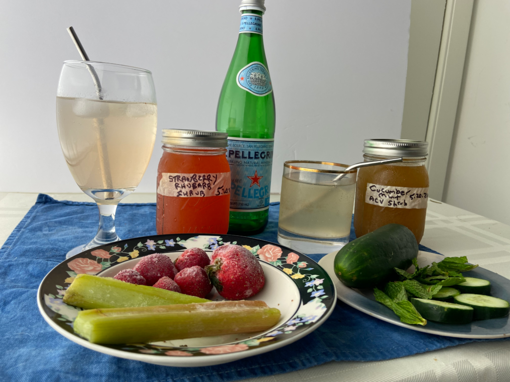 Photo example of what is a vinegar shrub showing strawberry rhubarb shrub and cucumber mint shrub in jars, mixed with sparkling water in glasses with metal straws, and the fresh fruits and veggies on plates in foreground.
