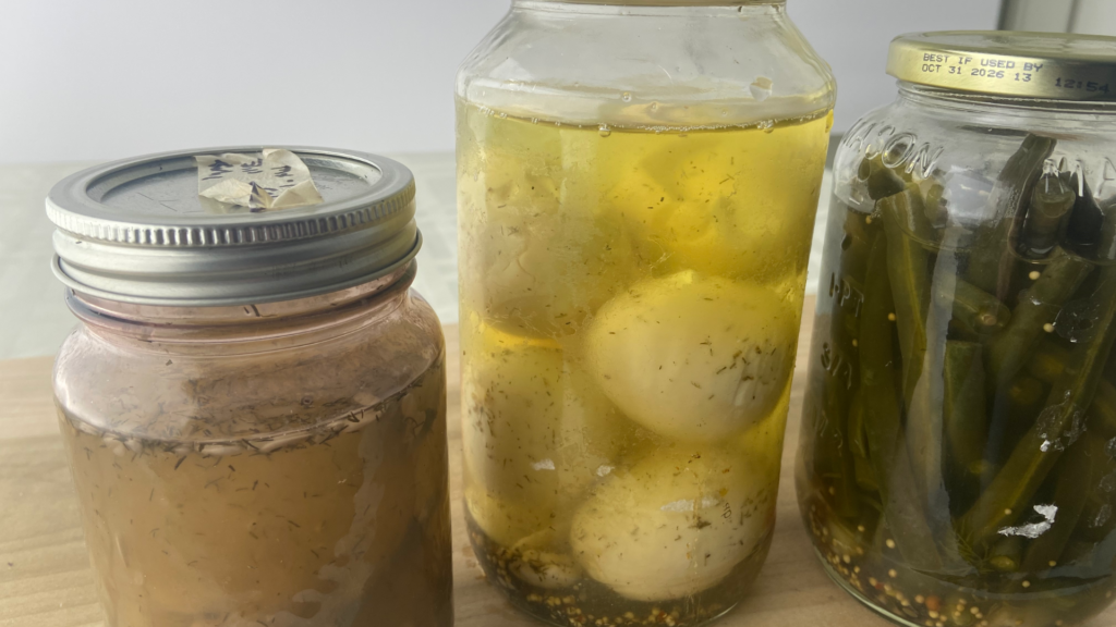 Jars of pickled cucumbers, pickled hard boiled eggs, and pickled string beans in brine with pickling spices visible
