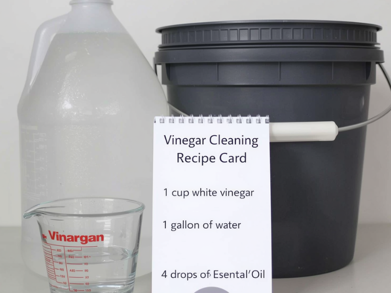 a straight on look at a jug of vinegar, a measuring cup, a bucket, and a Vinegar Cleaning Recipe Card that reads "1 cup white vinegar", 1 gallon of water, 4 drops of essential oil
