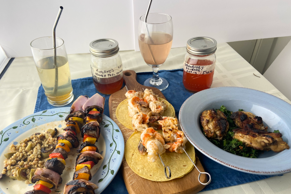 Display of vinegar summer mocktails made with summer shrubs and plates of grilled food in the foreground