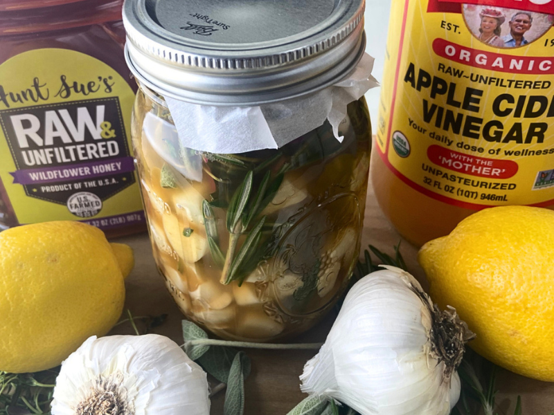closeup of a garlic oxymel fermenting in a jar with a container of raw honey and bottle of apple cider vinegar next to it and lemons and garlic in front