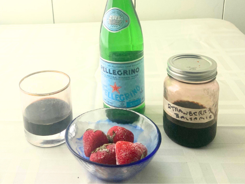a glass holding a balsamic vinegar strawberry shrub sits on a talbe with a bottle of pellegrino water, jar of strawberry balsamic shrub and bowl of fruit