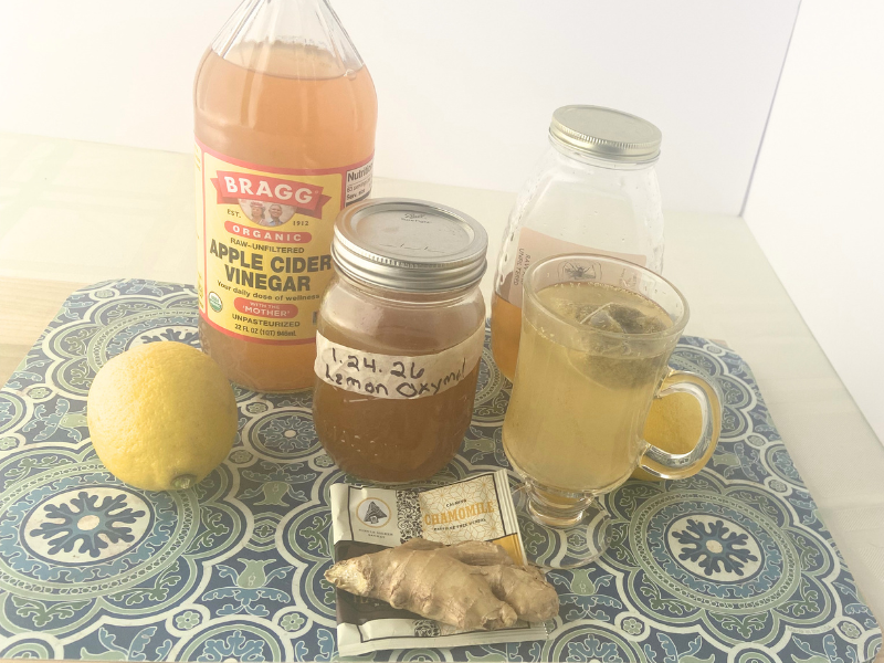 glass of camomile tea on mat with apple cider vinegar, ripe lemon, lemon oxymel, raw honey and ginger root.