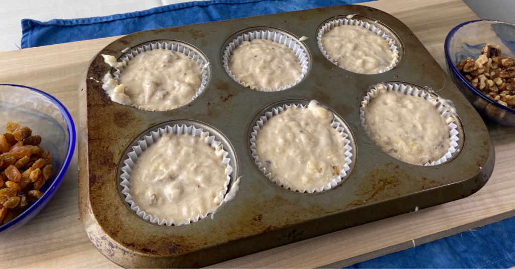 banana nut muffin batter in muffin pan with raisins in bowl on one side of pan and bowl with walnuts on the other side
