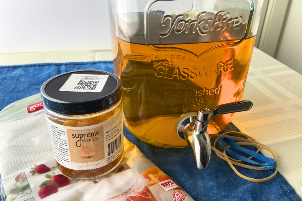 photo of decarbonated hard cider in a beverage dispenser, a jar of vinegar mother, cheesecloth, and rubber bands which are necessary to make apple cider vinegar from hard cider