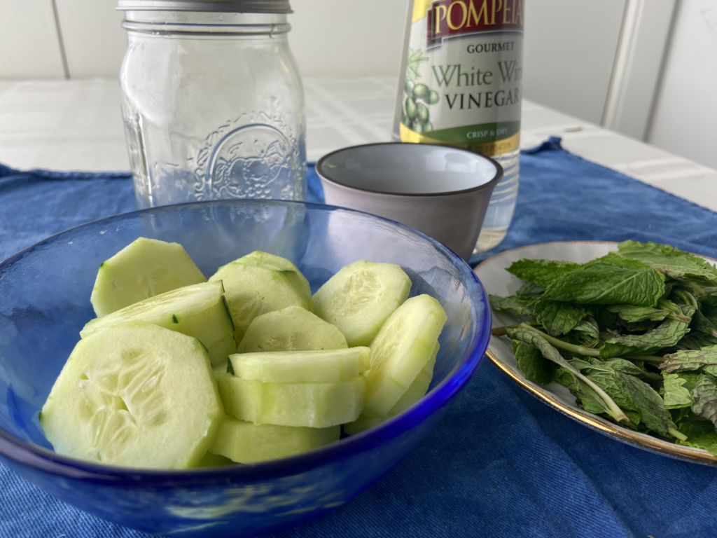 Closeup of ingredients for cucumber mint shrub that include sliced cucumber, fresh mint leaves, brown sugar and white wine vinegar and empty mason jar