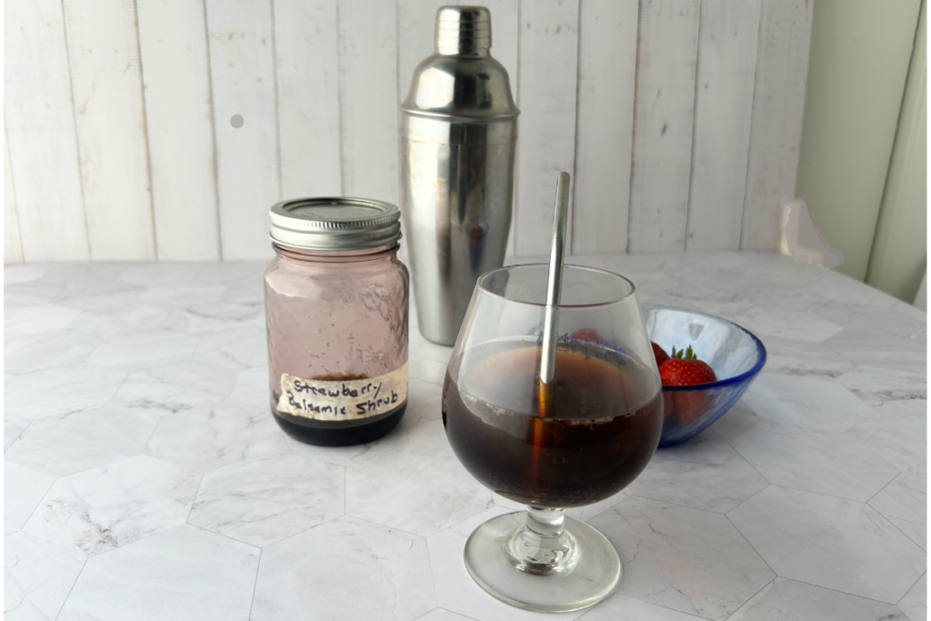 Strawberry  balsamic shrub in a jar as well as mixed in a glass with sparkling water and fruit and cocktail mixer in background