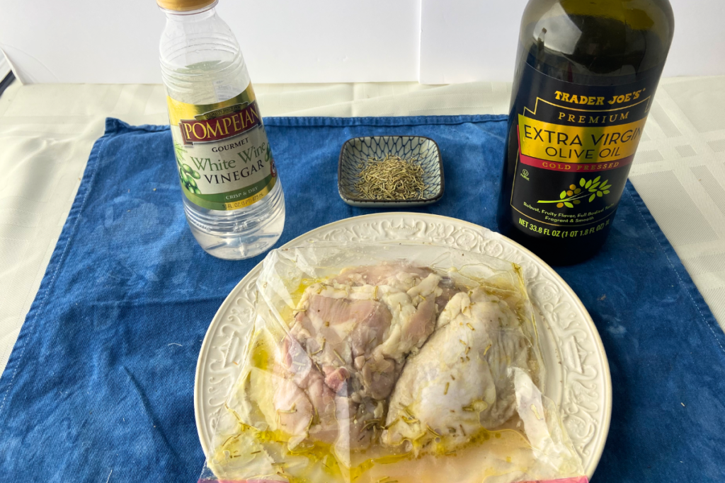 View of white wine vinegar, rosemary on a small plate, extra virgin olive oil, and chicken thighs marinating in a bag
