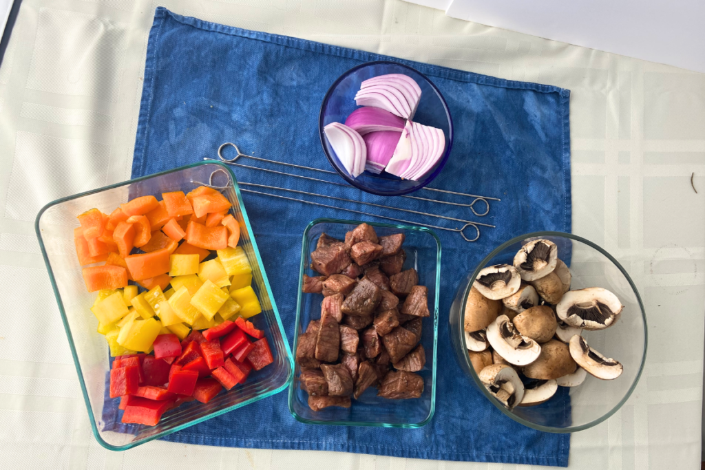 Overhead view of cut orange, yellow, and red bell peppers in a glass container, marinated beef cubes in a glass container, halved mushrooms in a glass bowl, quartered slices of red onion in a glass bowl and metal skewers.