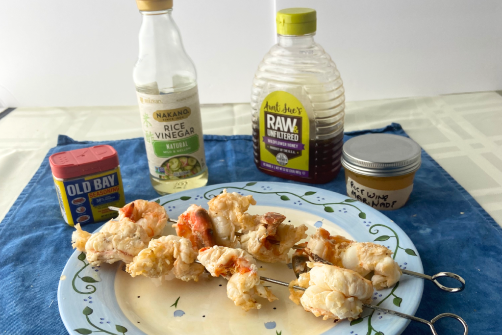Skewered marinated shrimp on a plate with Old Bay seasoning, rice wine vinegar, raw honey, and a small jar of rice wine marinade behind plate