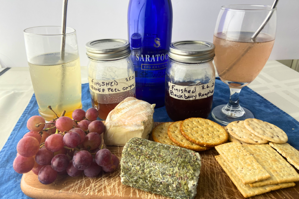 Food & Drink cheese board with red grapes, block of brie, log of chevre, crackers, oxymel mocktails and jars of oxymels and shrub