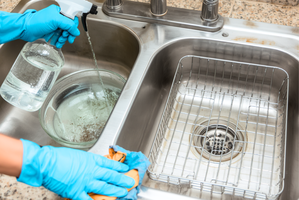 by gloved hands holding a spray bottle and a microfiber cloth to indicate Freshening a small kitchen sink and dish rack with a vinegar‑water mix.