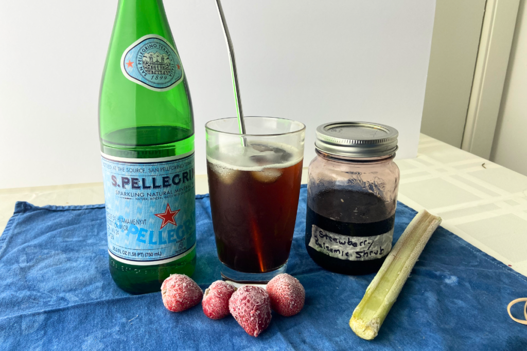 A bottle of sparkling water used to dilute a strawberry vinegar shrub in a glass with a metal straw and fruit arrayed alongside.