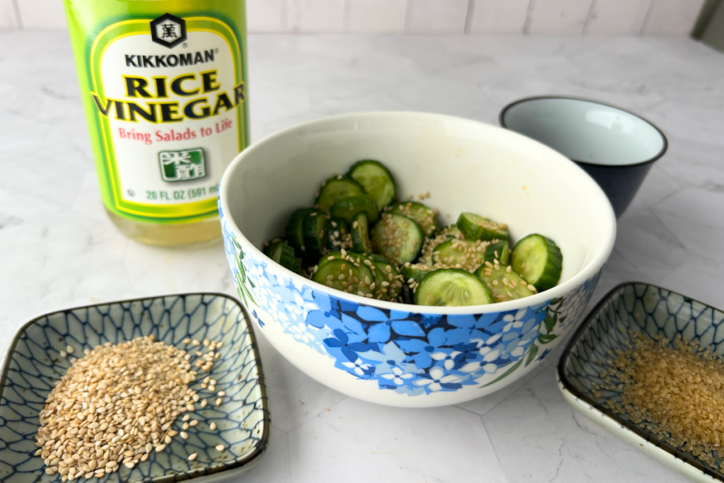 A Japanese cucumber salad in a bowl with a bottle of rice vinegar behind and sauce, sugar, and sesame seeds around the bowl
