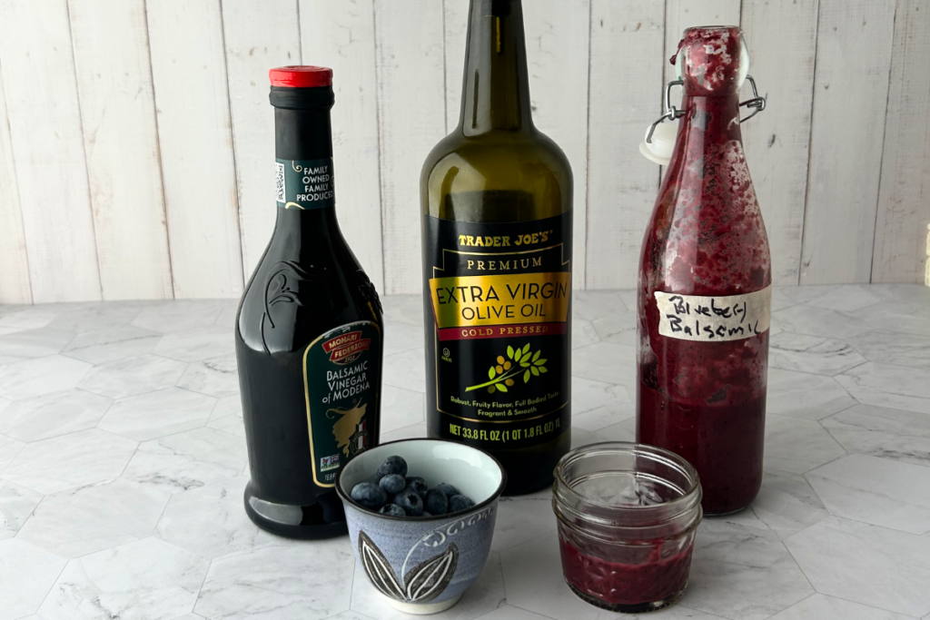 Line up of balsamic vinegar, olive oil, a bottle of DIY blueberry-balsamic vinaigrette and a small pouring cup