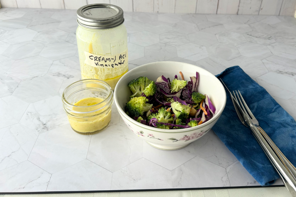 A jar of creamy ACV yogurt vinagrette with a chopped slaw salad and dinnerware