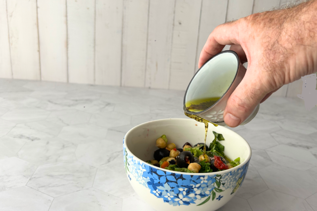 A hand pouring Mediterranean vinaigrette from cop over a mixed green salad with chickpeas and olives.