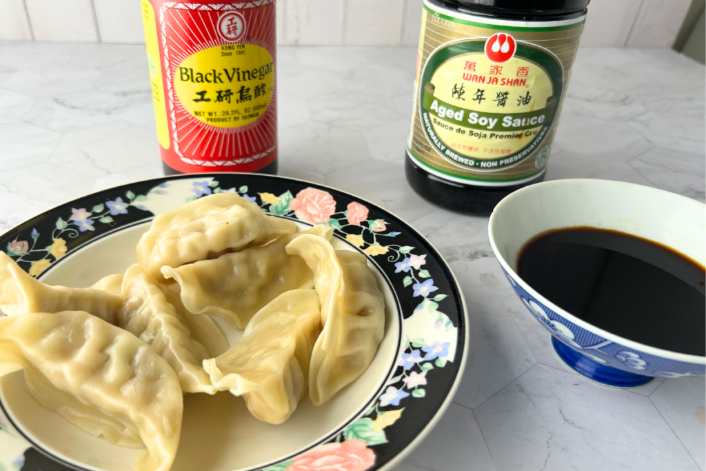 Steamed dumplings and small bowl of ponzu dipping sauce with bottle of black vinegar and soy sauce in background.