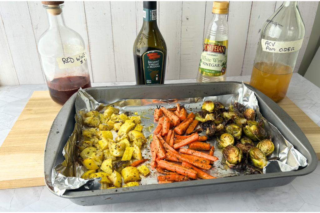 pan of roasted squash, roasted carrots, and roasted Brussel sprouts with bottles of red wine vinegar, balsamic vinegar, white wine vinegar, and apple cider vinegar in background.