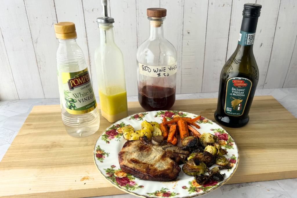 White wine vinegar, a white wine vinaigrette, red wine vinegar, and balsamic vinegar behind autumn vinegar health plate containing a vinegar-marinaded pork chop and roasted veggies with vinegar drizzles